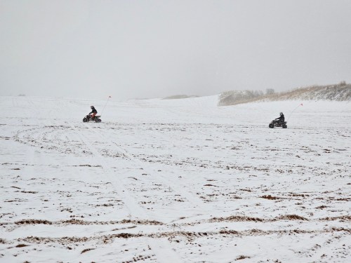 Two all-terrain vehicles ride across a snowy landscape under overcast skies.