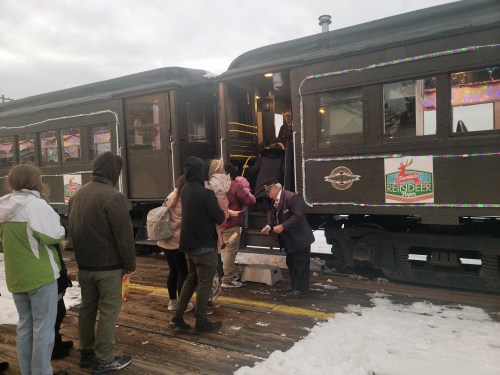 Passengers boarding a decorated train in a snowy setting, with a conductor assisting at the steps.