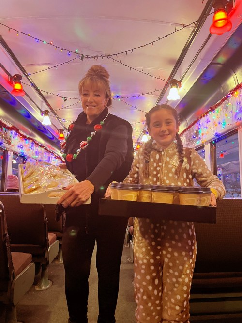 A woman and a girl in festive attire hold trays of cookies on a decorated train with colorful lights.