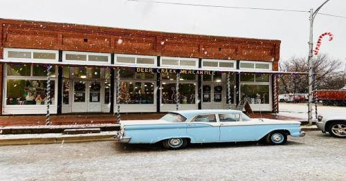 A vintage blue car parked in front of a festive mercantile store, with snow falling gently around.
