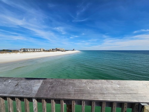 A serene beach view from a wooden pier, featuring clear blue water and a distant shoreline under a bright sky.
