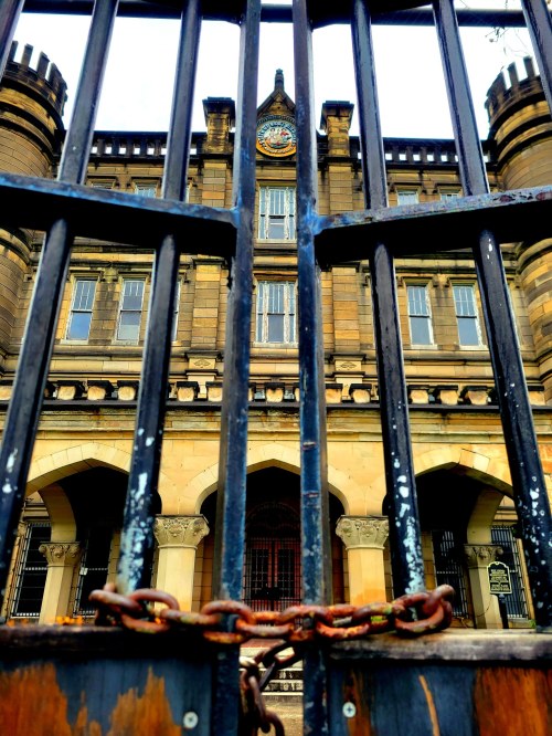A historic building viewed through a barred gate, with a clock tower and stone architecture visible in the background.