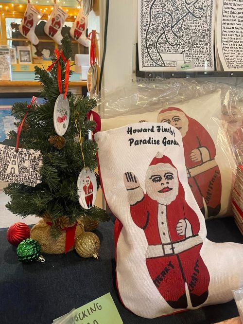 A small Christmas tree with ornaments next to decorative pillows featuring a Santa design and a sign reading "Howard Finley Paradise Garden."