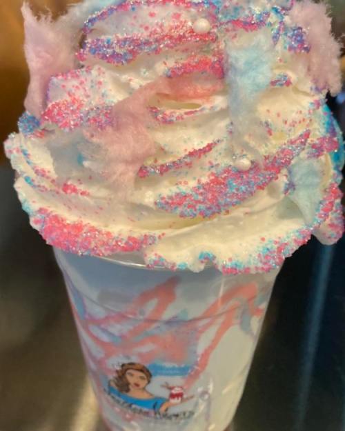 A colorful milkshake topped with whipped cream, pastel cotton candy, and sparkling sprinkles in a clear cup.