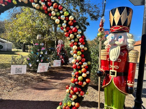 A festive scene featuring a nutcracker statue beside a decorated arch and Christmas trees in a sunny outdoor setting.