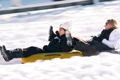 Two women joyfully sledding down a snowy hill, wearing winter clothing and laughing as they slide.