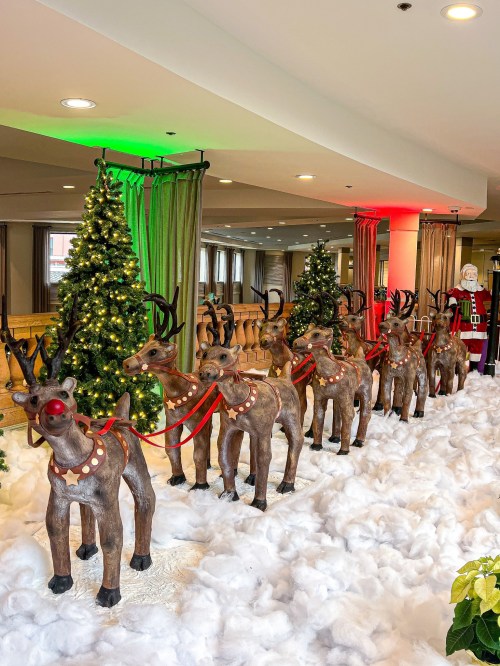 A festive display featuring reindeer with red harnesses, snow, Christmas trees, and a Santa figure in the background.