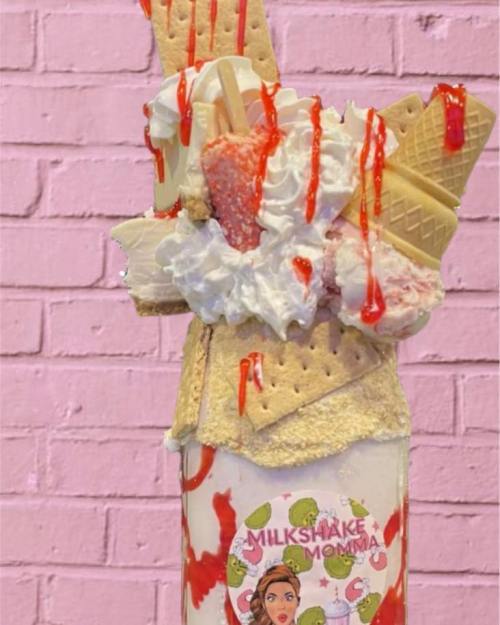 A tall milkshake topped with whipped cream, ice cream, strawberries, and drizzled with syrup, against a pink brick background.