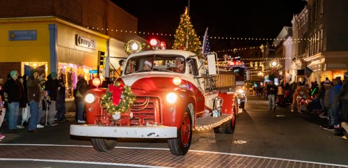 A vintage fire truck decorated for the holidays drives through a festive parade at night, surrounded by lights and spectators.