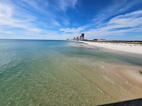 A serene beach scene with clear water, soft sand, and a skyline of buildings under a blue sky with wispy clouds.