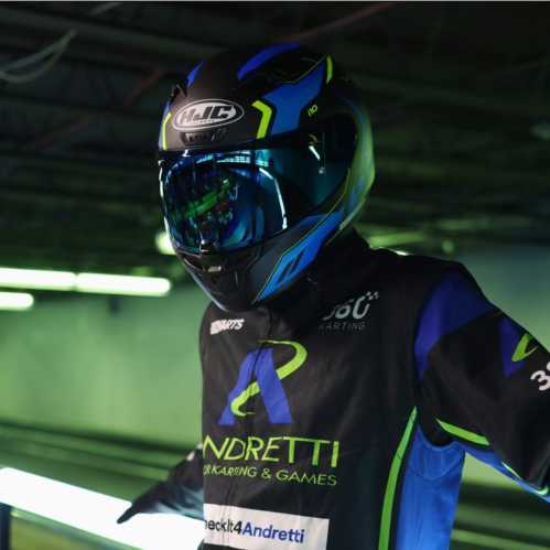 A go-kart racer in a blue and black helmet and racing suit stands in a dimly lit track area, ready to race.