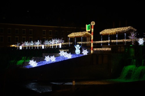Colorful holiday lights illuminate a snowy scene with a snowman and decorated trees by a water feature at night.