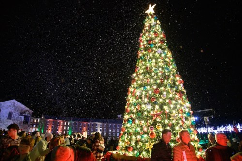 A large, decorated Christmas tree glows brightly in a snowy night, surrounded by a crowd of festive people.