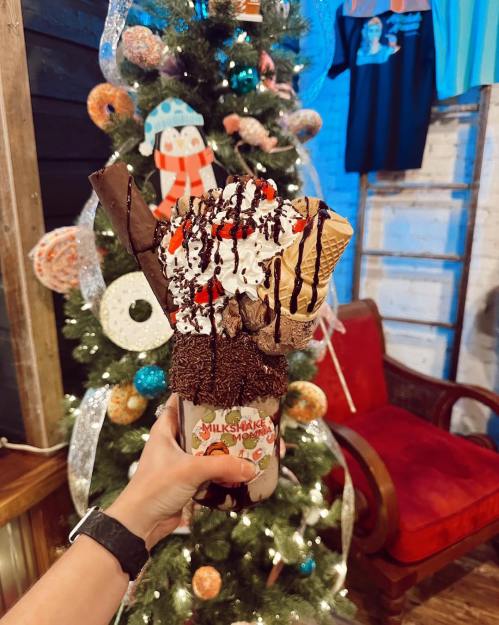 A festive milkshake topped with whipped cream, chocolate, and candy, held in front of a decorated Christmas tree.