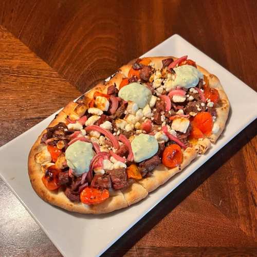 A flatbread pizza topped with grilled meat, cherry tomatoes, pickled onions, and dollops of green sauce on a white plate.