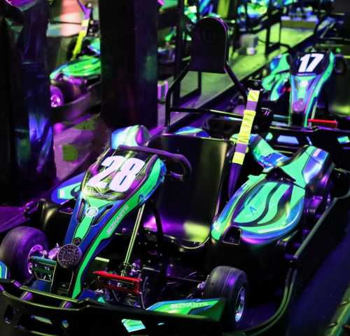 A row of colorful go-karts with vibrant green and blue designs, illuminated by neon lights in a racing venue.