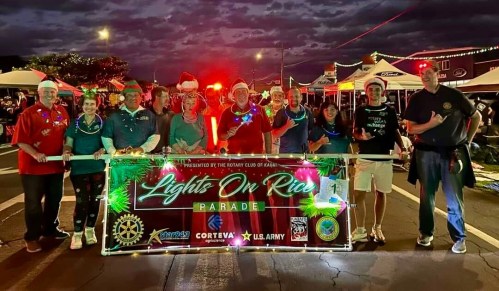 A group of people in festive attire holding a "Lights On Rice Parade" banner, with holiday lights and decorations.
