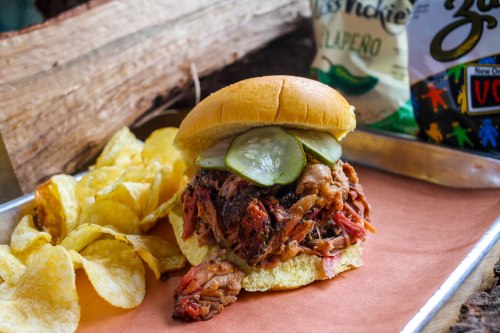 A pulled pork sandwich topped with pickles, served with potato chips on the side, on a rustic wooden background.