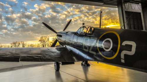 A vintage aircraft with a propeller, parked in a hangar, against a backdrop of a colorful sunset sky.