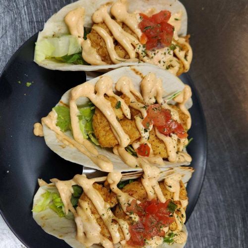 Three tacos filled with lettuce, crispy chicken, pico de gallo, and drizzled with a creamy sauce on a black plate.