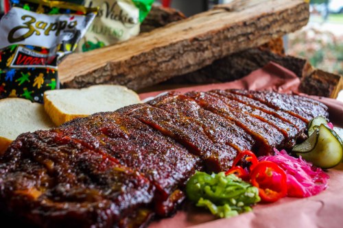 A platter of smoked ribs with pickles, jalapeños, pink onions, and slices of bread, set on a rustic wooden background.