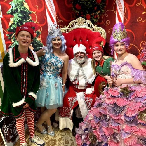 A group of festive characters, including Santa, pose together in a colorful holiday setting.