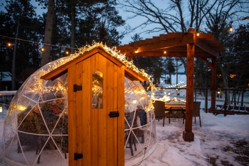 A cozy igloo-shaped structure with fairy lights, surrounded by snow and trees, near a wooden pergola with string lights.
