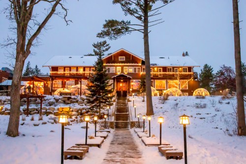 A cozy lodge adorned with lights, surrounded by snow and trees, with a pathway lined by lanterns leading to the entrance.
