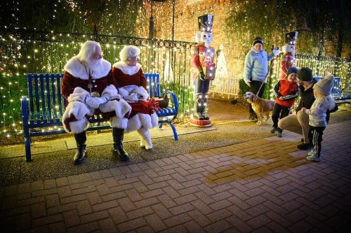 A festive scene with Santa and Mrs. Claus sitting on a bench, surrounded by holiday lights and families enjoying the decorations.
