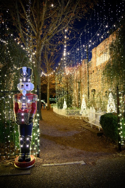 A festive pathway lined with lights, featuring a nutcracker statue and decorated trees in the background.