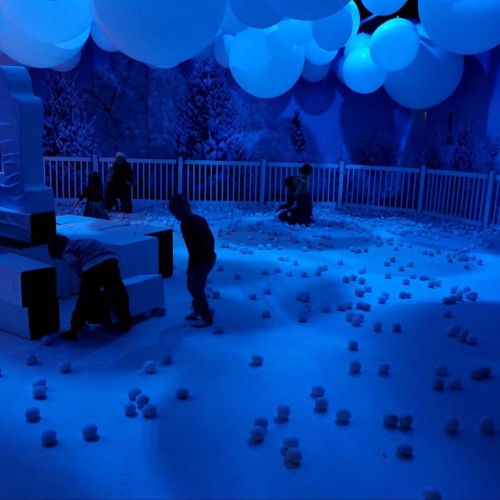 Children playing in a blue-lit room filled with soft snowballs and large white balloons, resembling a winter scene.