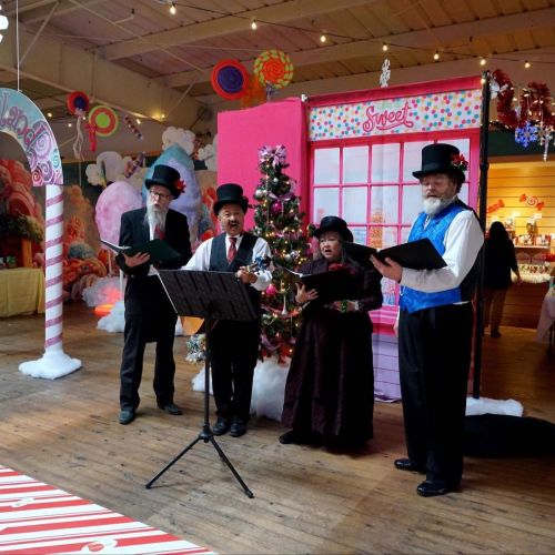 Four carolers in festive attire sing in front of a decorated Christmas backdrop with a tree and colorful decorations.