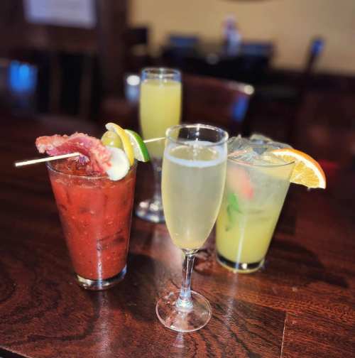 Four colorful cocktails on a wooden table, including a Bloody Mary, mimosa, and two fruity drinks with garnishes.