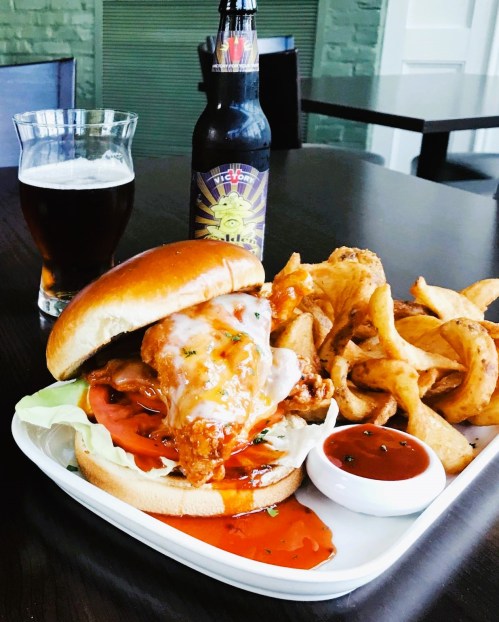 A plate featuring a spicy chicken sandwich with lettuce and tomato, served with curly fries and a beer.