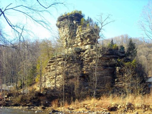 A tall rock formation surrounded by trees, with a clear blue sky in the background and a river in the foreground.