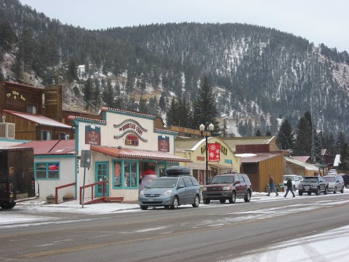 A snowy street scene with colorful storefronts and vehicles parked along the road, set against a mountainous backdrop.