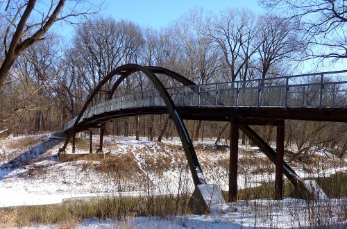 A curved pedestrian bridge spans a snowy landscape, surrounded by bare trees and a calm river below.
