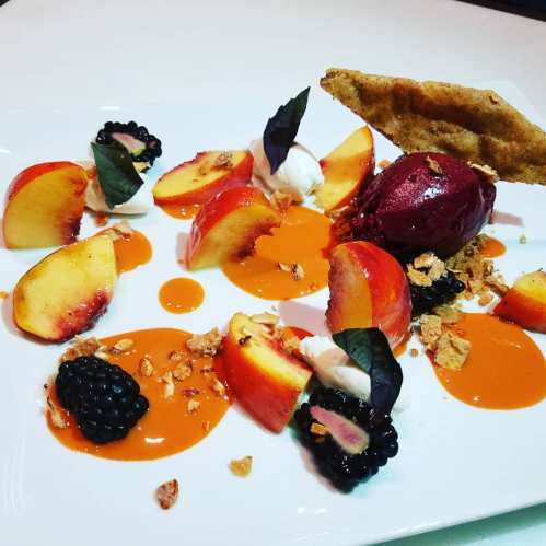 A colorful dessert plate featuring sliced peaches, blackberries, sorbet, and garnishes on a white background.