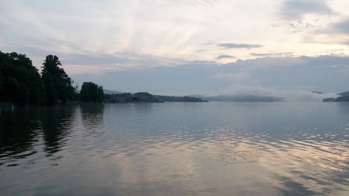 A serene lake at dawn, with mist rising from the water and soft clouds reflecting in the calm surface.