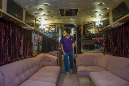 A young man stands inside a vintage bus, featuring plush seating and dim lighting, with decorative walls and curtains.