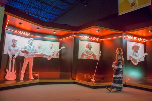 A visitor admires white sculptures of musicians Alabama, Lionel Richie, and Hank Williams Jr. in a dimly lit exhibit.