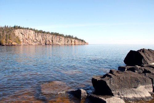 A serene lake scene with calm waters, rocky shore, and a forested cliff in the background under a clear blue sky.