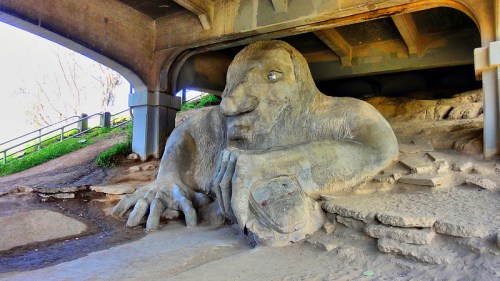 A large stone troll sculpture rests under a bridge, with a hand on the ground and a thoughtful expression.