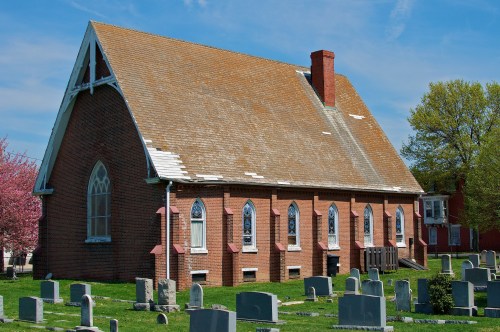 A brick church with a steep roof, surrounded by a cemetery and blooming trees under a clear blue sky.