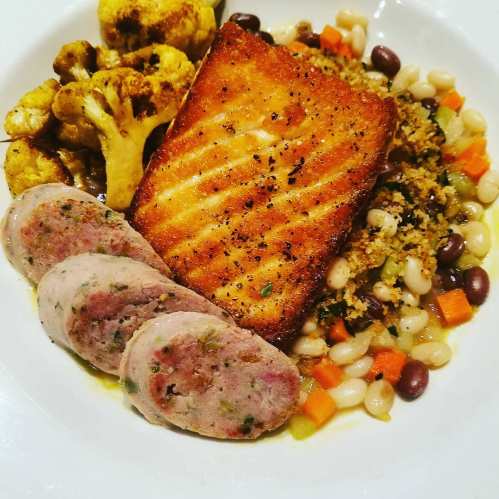 A plate featuring grilled salmon, sausage slices, mixed beans, couscous, and roasted cauliflower.