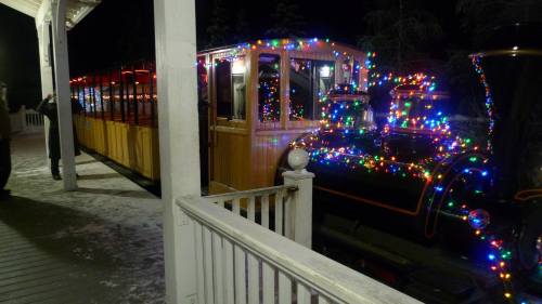 A brightly lit train decorated with colorful Christmas lights, parked beside a white wooden structure in a snowy setting.