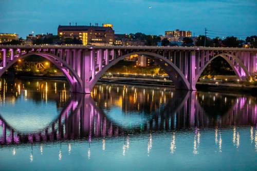 A bridge with purple lights arches over a calm river, reflecting the colorful lights and nearby buildings at dusk.