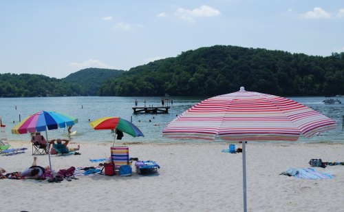 Colorful umbrellas shade beachgoers on a sandy shore by a calm lake, surrounded by green hills under a clear blue sky.