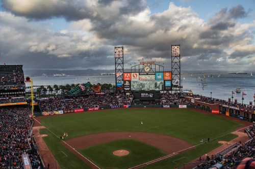 A panoramic view of a baseball stadium by the water, with fans in the stands and boats in the background under a cloudy sky.