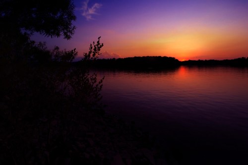 A serene sunset over a calm lake, with silhouettes of trees and rocks in the foreground against a colorful sky.